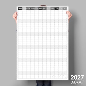 May include: A large, white 2027 year planner with a grid layout, held by a person. The months are listed across the top in a dark font. The size is A0/A1.
