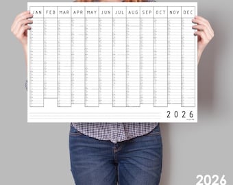 May include: A white 2026 year planner calendar with monthly columns and daily rows. The calendar is held by a person wearing a plaid shirt and jeans. The calendar has the text "2026" at the bottom.