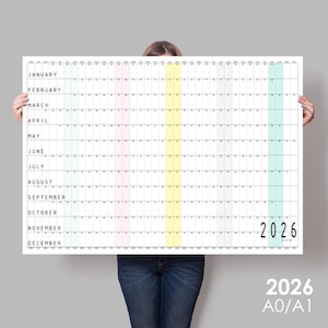 May include: A large white 2026 year planner with a grid layout, featuring months and dates. Vertical stripes in pink, yellow, and green highlight specific sections. The year "2026" is printed in black. The planner is held by a person.