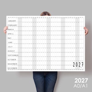 May include: A large white 2027 year planner with a grid layout, featuring months and days. The calendar is held up by a person. The text "2027" is visible in the bottom right corner. The size is A0/A1.