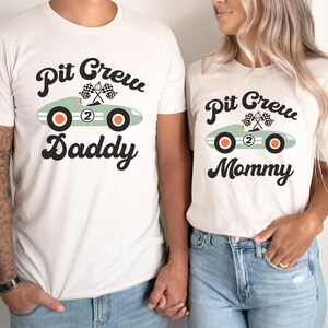 Pu&ograve; includere: Due magliette bianche con una grafica di un'auto da corsa verde e il testo "Pit Crew Daddy" e "Pit Crew Mommy".