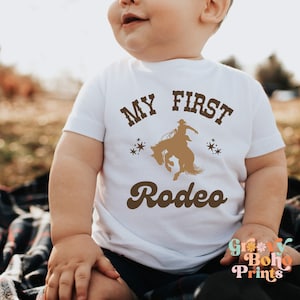 May include: A white baby onesie with brown text that reads "My First Rodeo" and a silhouette of a cowboy riding a bucking bronco. The onesie has a retro font style.