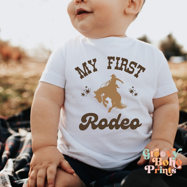 May include: A white baby onesie with brown text that reads "My First Rodeo" and a silhouette of a cowboy riding a bucking bronco. The onesie has a retro font style.