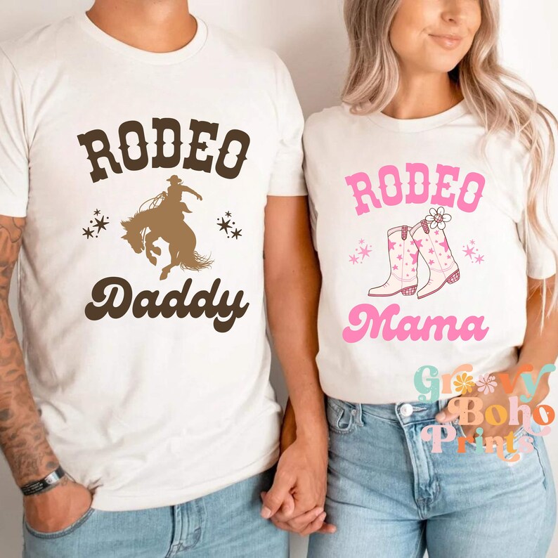 May include: Two white t-shirts with the text "Rodeo Daddy" and "Rodeo Mama" in brown and pink lettering. The "Daddy" shirt features a silhouette of a cowboy riding a bucking bronco. The "Mama" shirt features a pair of pink cowboy boots with stars and a flower.