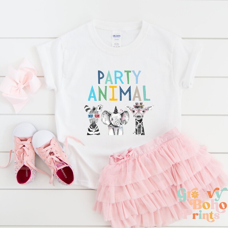 Puede incluir: Camiseta blanca con el texto "PARTY ANIMAL" en letras de colores. Tres animales de dibujos animados con sombreros de fiesta y accesorios se muestran debajo del texto. La camiseta est&aacute; sobre una superficie blanca con una falda de tut&uacute; rosa, un lazo rosa y zapatillas rosas.