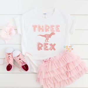 以下が含まれることがあります： 「THREE REX」の文字とピンクの恐竜のグラフィックが入った白いTシャツ。恐竜には数字の「3」が入っています。ピンクのチュチュ、ピンクの靴、ピンクのリボンも画像にあります。このシャツは子供の3歳の誕生日用です。