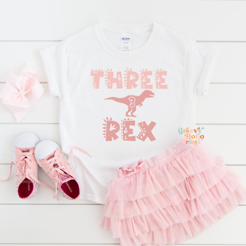 以下が含まれることがあります： 「THREE REX」の文字とピンクの恐竜のグラフィックが入った白いTシャツ。恐竜には数字の「3」が入っています。ピンクのチュチュ、ピンクの靴、ピンクのリボンも画像にあります。このシャツは子供の3歳の誕生日用です。