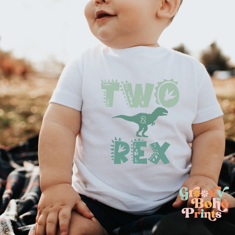 K&ouml;nnte beinhalten: Ein wei&szlig;es T-Shirt f&uuml;r Kleinkinder mit einem gr&uuml;nen Dinosaurier-Motiv und dem Text "TWO REX" mit der Zahl "2" im Dinosaurier.