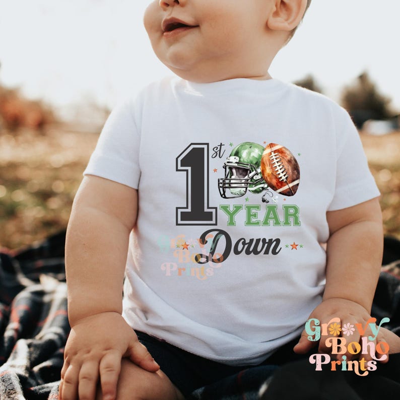 May include: White toddler t-shirt with a green and black football helmet and football graphic. The text on the shirt reads "1st Year Down".