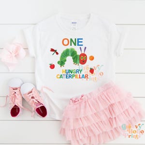 May include: White t-shirt with a colorful graphic of a green caterpillar, an apple, and the words "ONE HUNGRY CATERPILLAR." A pink tutu skirt and pink sneakers are next to the shirt. The shirt is a Gildan brand.