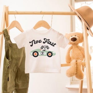 Pu&ograve; includere: Maglietta bianca con un'immagine di un'auto da corsa verde e il testo "Two Fast" in nero. L'auto ha il numero "2" sul lato e bandiere a scacchi.