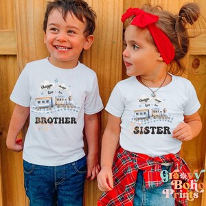 May include: Two children wearing white t-shirts with a blue train design and the words "chugga chugga choo choo" and "brother" and "sister".