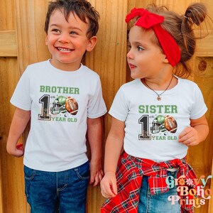 May include: Two children wearing white t-shirts with green and brown football helmet and ball graphics. The shirts say "Brother of the 1 Year Old" and "Sister of the 1 Year Old".