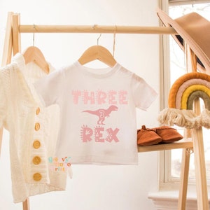以下が含まれることがあります： ピンク色の「THREE REX」の文字と恐竜のグラフィックがプリントされた白い幼児用Tシャツ。木製のラックに他の衣類やアクセサリーと一緒に吊るされています。
