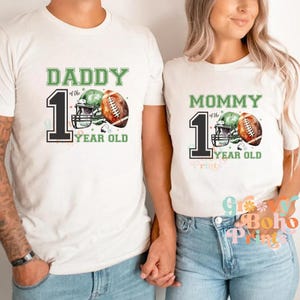 May include: White t-shirts with green and black text that reads "Daddy of the 1 Year Old" and "Mommy of the 1 Year Old".  Each shirt features a football helmet and football with watercolor-style green and brown accents.