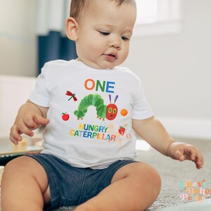 May include: A white short-sleeved t-shirt featuring a colorful graphic of a green caterpillar, an apple, an orange, a strawberry, and a dragonfly. The word "ONE" is above the caterpillar, and "HUNGRY CATERPILLAR" is below. The child is wearing blue shorts.