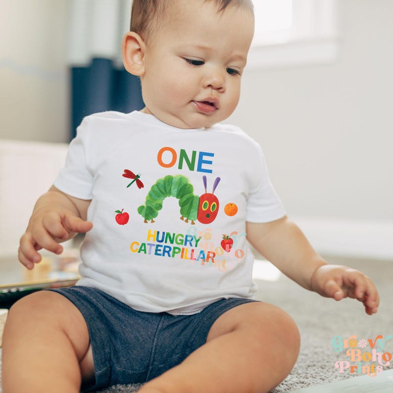 May include: A white short-sleeved t-shirt featuring a colorful graphic of a green caterpillar, an apple, an orange, a strawberry, and a dragonfly. The word "ONE" is above the caterpillar, and "HUNGRY CATERPILLAR" is below. The child is wearing blue shorts.