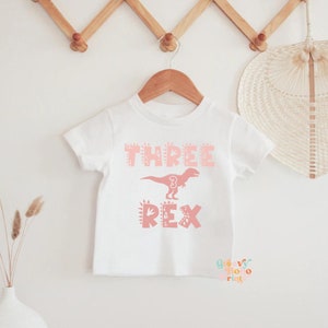 以下が含まれることがあります： ピンクの恐竜のグラフィックと「THREE REX」の文字が入った白いTシャツ。数字の「3」は恐竜のデザインに組み込まれています。木製のハンガーにTシャツが掛けられています。