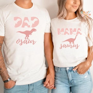 以下が含まれることがあります： ピンクの恐竜グラフィックがプリントされた、お揃いのクリーム色のTシャツ2枚。1枚は「DAD osaur」とT-Rexのシルエット、もう1枚は「MAMA saurus」とブロントサウルスのシルエット。Tシャツはブルージーンズと合わせて着用されています。