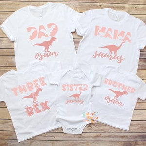 以下が含まれることがあります： ピンク色の恐竜をテーマにしたテキストがプリントされた白いTシャツとボディスーツ。Tシャツには「DAD osaur」、「MAMA saurus」、「THREE REX」、「SISTER saurus」、「BROTHER osaur」と書かれています。恐竜のシルエットが特徴です。