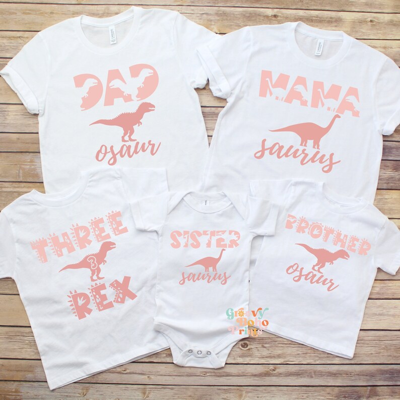 以下が含まれることがあります： ピンク色の恐竜をテーマにしたテキストがプリントされた白いTシャツとボディスーツ。Tシャツには「DAD osaur」、「MAMA saurus」、「THREE REX」、「SISTER saurus」、「BROTHER osaur」と書かれています。恐竜のシルエットが特徴です。