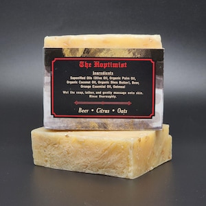 May include: Two bars of handmade soap, light beige in colour. The top bar has a black label with the text "The Hoptimist" and ingredient information. Ingredients include beer, citrus, and oats. The soap is on a dark background.