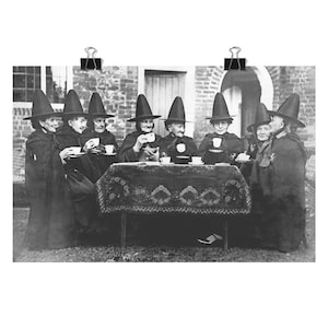 Könnte beinhalten: Ein Schwarzweißfoto von acht Frauen, die schwarze spitze Hüte und Umhänge tragen und um einen Tisch mit Tassen und Untertassen sitzen. Sie trinken Tee im Freien vor einem Backsteingebäude.