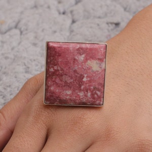 May include: A square-shaped ring featuring a large, pink gemstone with a marbled appearance. The gemstone is set in a silver-colored metal band. The ring is a statement piece, showcasing a bold and eye-catching design.