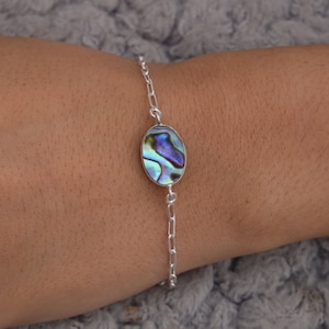 May include: A silver chain bracelet with an oval-shaped abalone shell pendant. The shell has iridescent blue, green, and purple colors.