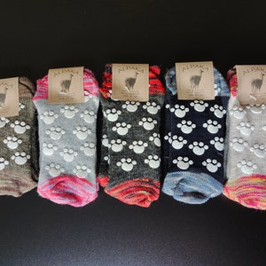 May include: Five pairs of alpaca wool socks with grippy paw prints on the bottom. The socks are in various colors, including gray, brown, black, and white. The socks have a tag that says "Alpaca" on them.