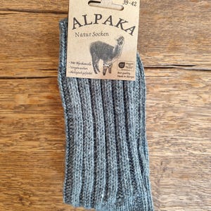 May include: A pair of gray Alpaka Natur Socken, size 39-42, with a ribbed knit design. The socks have a tag with an alpaca illustration and text in German. The socks are made of alpaca wool and are pre-washed and ecologically dyed.