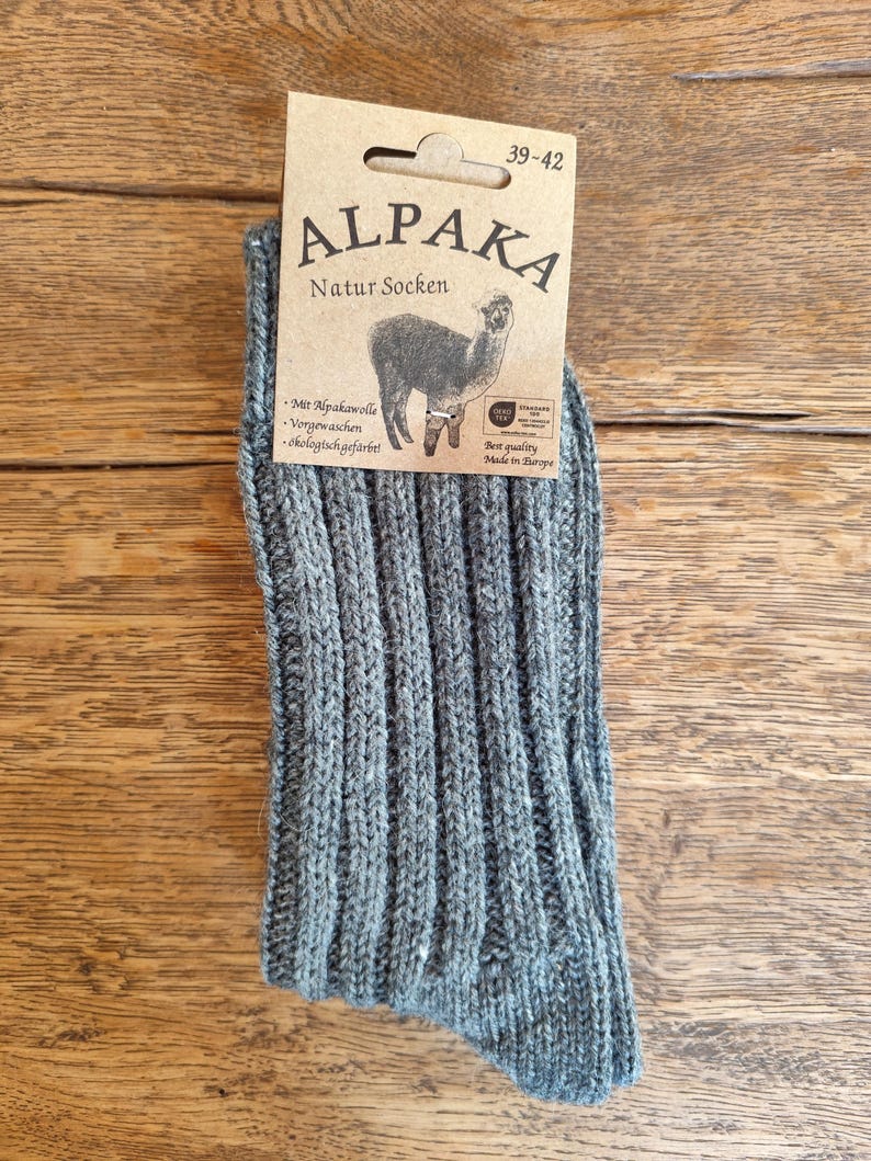 May include: A pair of gray Alpaka Natur Socken, size 39-42, with a ribbed knit design. The socks have a tag with an alpaca illustration and text in German. The socks are made of alpaca wool and are pre-washed and ecologically dyed.