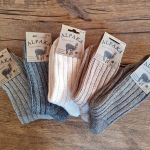 May include: Five pairs of ribbed knit socks in various neutral colors, including gray, beige, and off-white, are displayed on a wooden surface. Each pair has a tag that reads "ALPAKA" with an illustration of an alpaca.