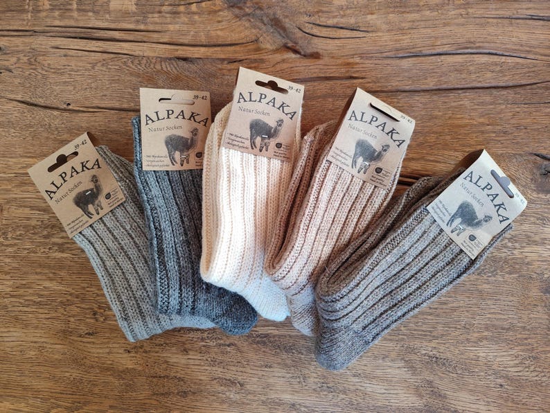 May include: Five pairs of ribbed knit socks in various neutral colors, including gray, beige, and off-white, are displayed on a wooden surface. Each pair has a tag that reads "ALPAKA" with an illustration of an alpaca.