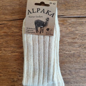 May include: Cream-colored Alpaka Natur Socken socks, size 39-42, with a ribbed knit design. The packaging features a brown label with the word "ALPAKA" and an illustration of an alpaca. Made in Europe.