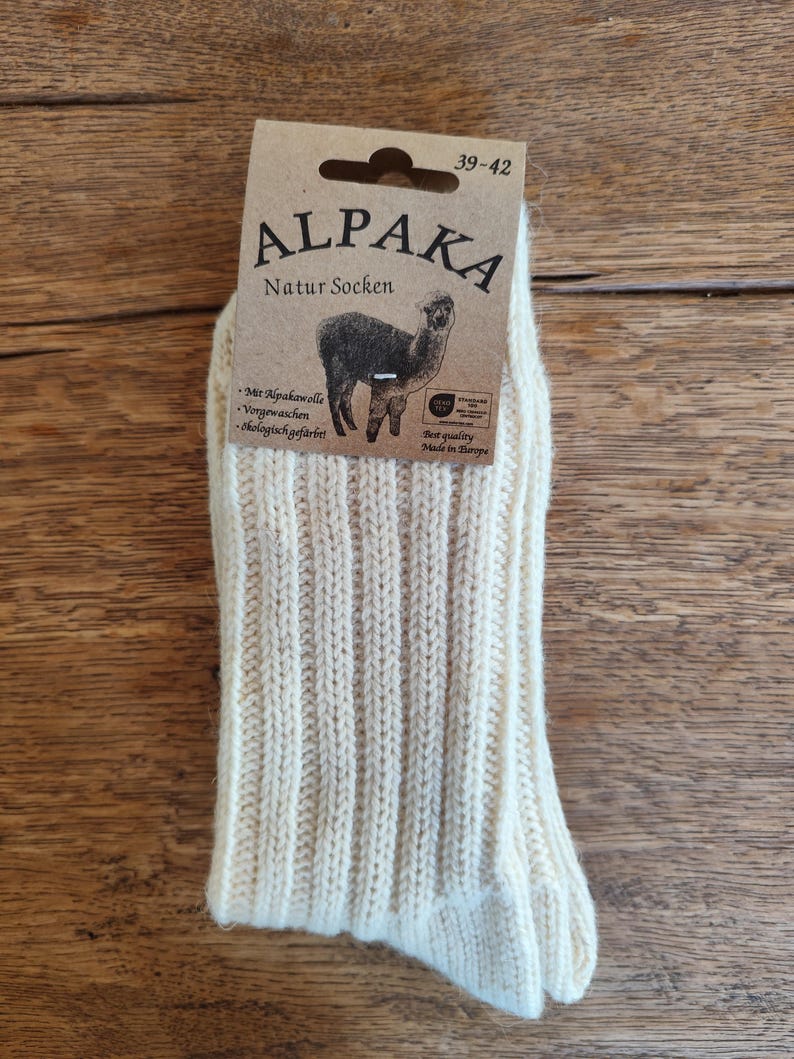 May include: Cream-colored Alpaka Natur Socken socks, size 39-42, with a ribbed knit design. The packaging features a brown label with the word "ALPAKA" and an illustration of an alpaca. Made in Europe.