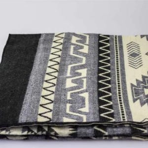 May include: Folded blanket with a geometric pattern in shades of black, gray, and cream. The blanket features a dark gray border and a repeating design of zigzags, diamonds, and other shapes. The blanket is made of a soft material.
