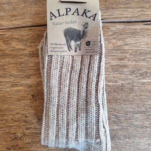 May include: A pair of beige Alpaka Natur Socken socks with a ribbed knit design. The socks have a tag with the text "ALPAKA Natur Socken" and an illustration of an alpaca. The tag also includes the size range 39-42.