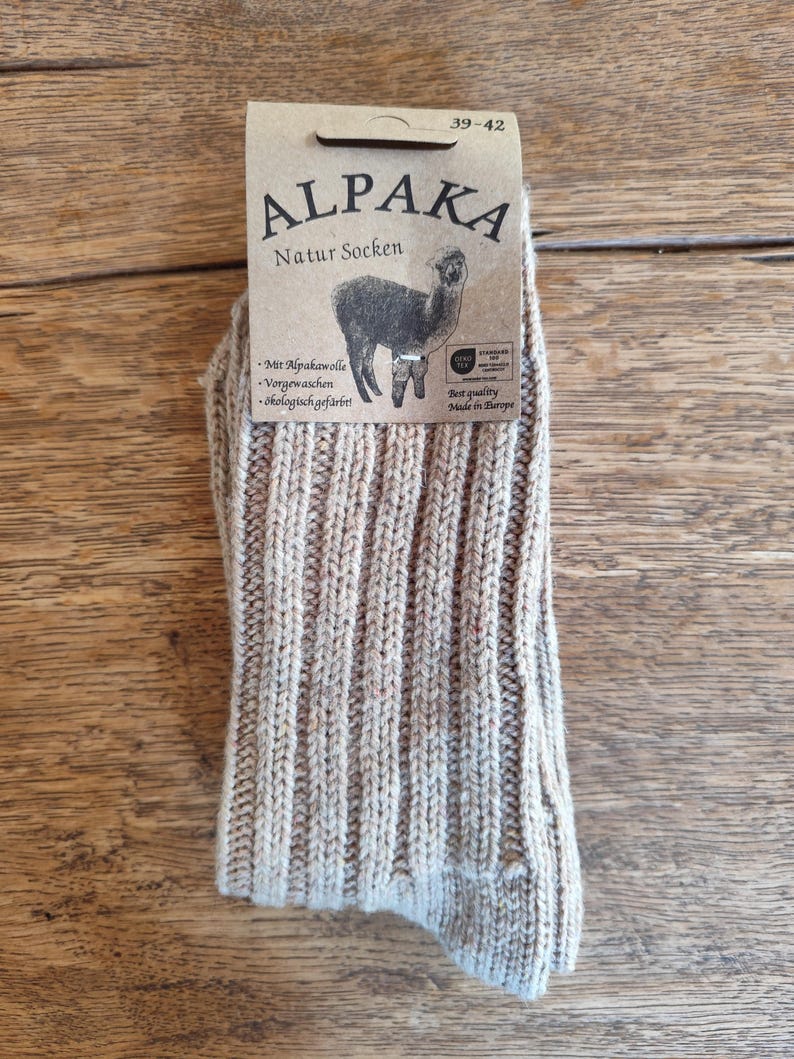 May include: A pair of beige Alpaka Natur Socken socks with a ribbed knit design. The socks have a tag with the text "ALPAKA Natur Socken" and an illustration of an alpaca. The tag also includes the size range 39-42.