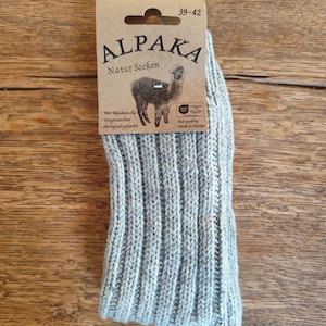 May include: A pair of light gray Alpaka Natur Socken socks with a ribbed knit design. The socks are packaged with a cardboard tag featuring the brand name, an alpaca illustration, and text in German. The tag indicates sizes 39-42.