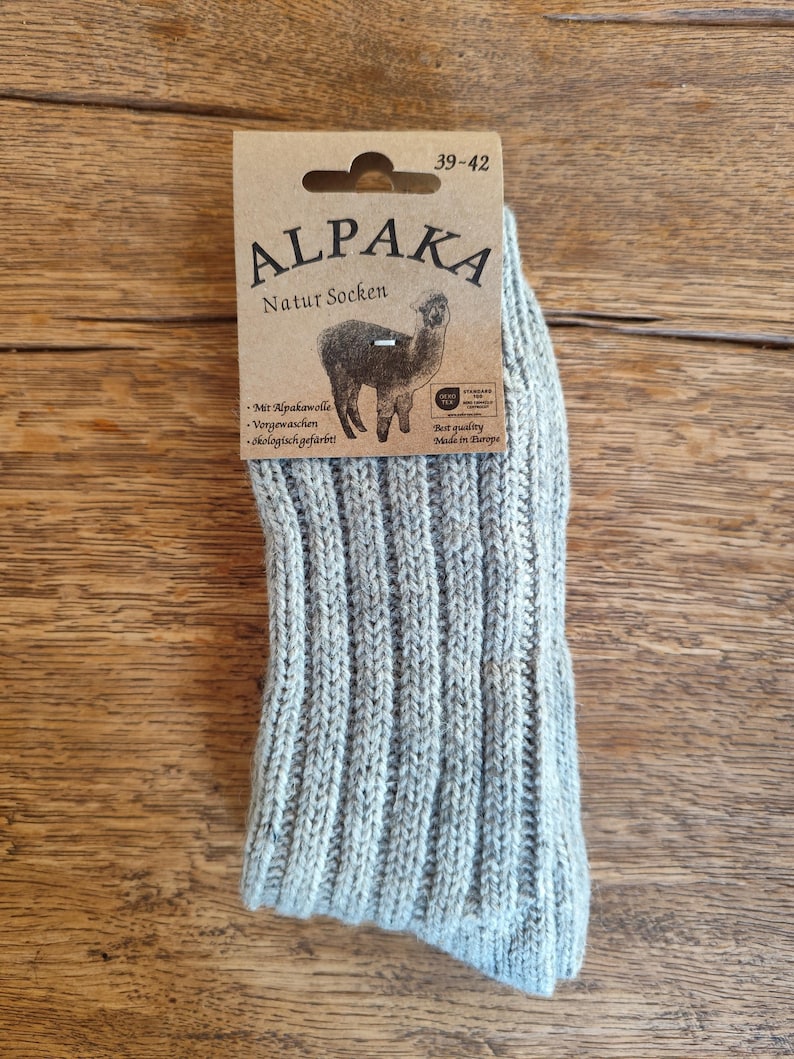 May include: A pair of light gray Alpaka Natur Socken socks with a ribbed knit design. The socks are packaged with a cardboard tag featuring the brand name, an alpaca illustration, and text in German. The tag indicates sizes 39-42.