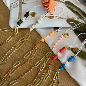 Puede incluir: Un conjunto de seis colgantes para teléfono con cadenas doradas y cuentas de colores. Cada colgante tiene un charm en forma de corazón con la palabra "Love" escrita en él. Los colgantes están dispuestos en una fila sobre una superficie de madera.