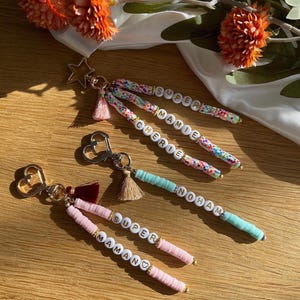 May include: Keyrings with beaded accents and the words "SUPER", "MAMIE", "CHERIE", and "NOHAM" spelled out in white letter beads. Each keyring has a different charm, including a star, heart, and tassel.