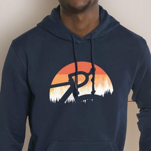 Peut inclure: Sweat à capuche bleu marine avec une poche avant et une capuche à cordon. Le graphisme sur le devant représente une silhouette de guidon de vélo sur un coucher de soleil aux teintes orange, rouge et blanc, et un contour de forêt.
