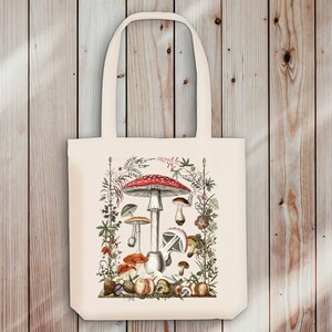May include: A white tote bag with a colorful illustration of mushrooms and foliage. The mushrooms are red, white, and brown with a variety of shapes and sizes. The foliage is green and brown with small flowers.