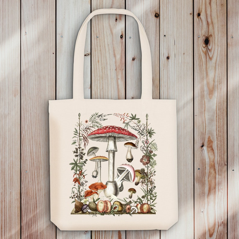 May include: A white tote bag with a colorful illustration of mushrooms and foliage. The mushrooms are red, white, and brown with a variety of shapes and sizes. The foliage is green and brown with small flowers.