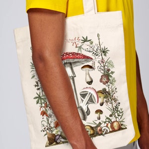 May include: A white canvas tote bag with a colorful illustration of mushrooms and flowers. The bag has a yellow strap and is perfect for carrying groceries or other items.