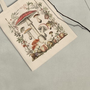May include: A white tote bag with a vintage illustration of mushrooms and other botanical elements. The mushrooms are red and white with brown stems. The background is a cream color.
