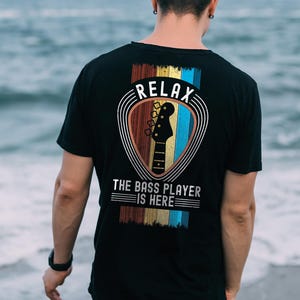 Peut inclure: T-shirt noir avec un motif graphique au dos. Le motif représente une silhouette de basse dans un bouclier, avec le mot "RELAX" au-dessus et "THE BASS PLAYER IS HERE" en dessous. Le design utilise des rayures rouges, dorées, bleues et marron.