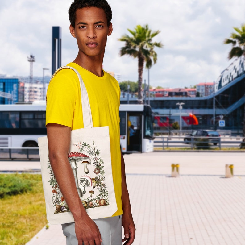 May include: A person is carrying a white tote bag with a vintage illustration of mushrooms and greenery. The bag has a brown handle and is made of canvas.